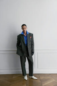 Man in a gray double-breasted suit with a blue striped shirt, pink pocket square, white shoes, and black gloves stands against a white paneled wall.