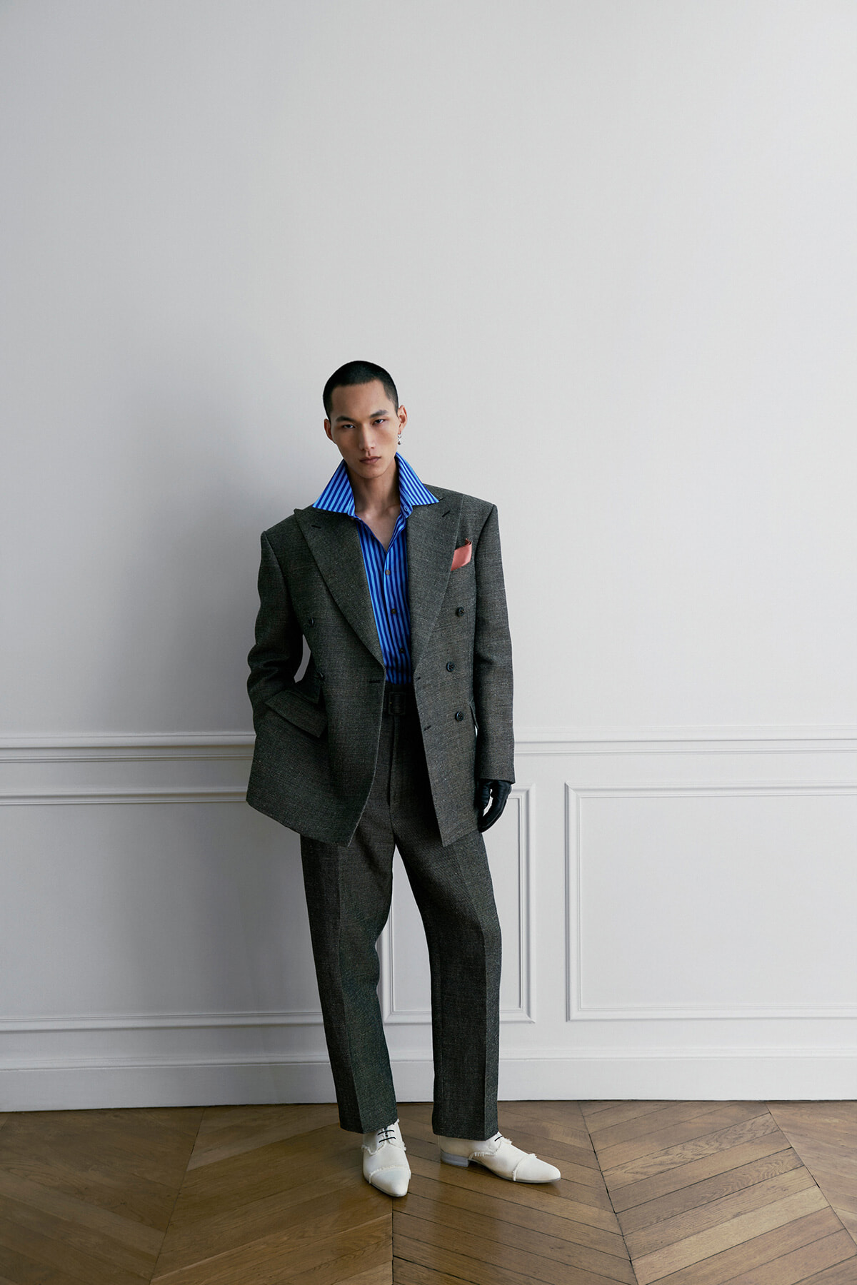 Man in a gray double-breasted suit with a blue striped shirt, pink pocket square, white shoes, and black gloves stands against a white paneled wall.