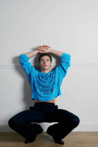 Young man in a bright blue sweater crouches in a wide stance with arms raised and hands clasped over his head against a plain white wall of a studio.