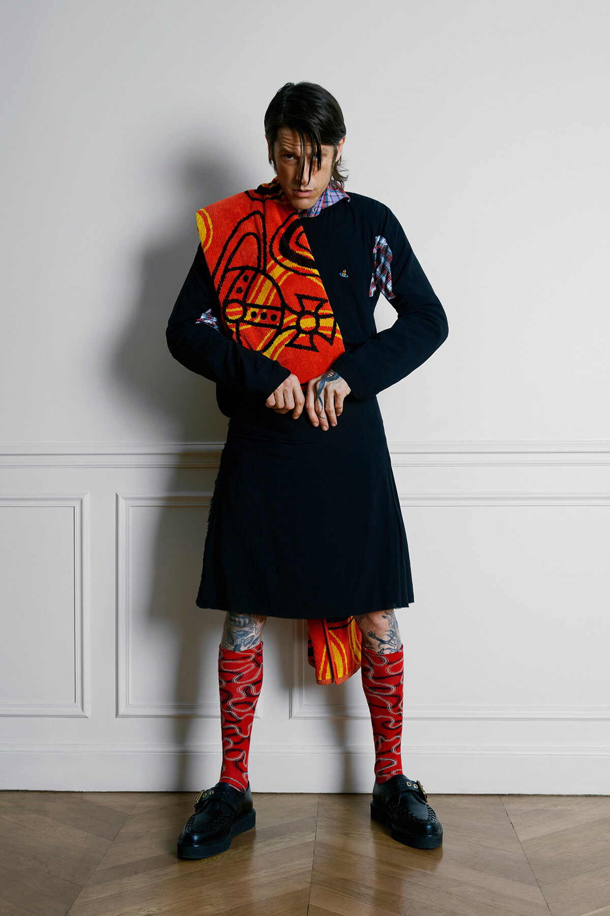 Person wearing a black tunic dress with red patterned socks, holding a bold red textile draped over one shoulder against a white wall.