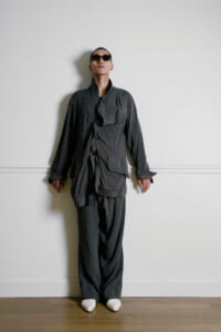 Person wearing an oversized dark gray suit and sunglasses, standing against a white wall with arms relaxed at the sides.