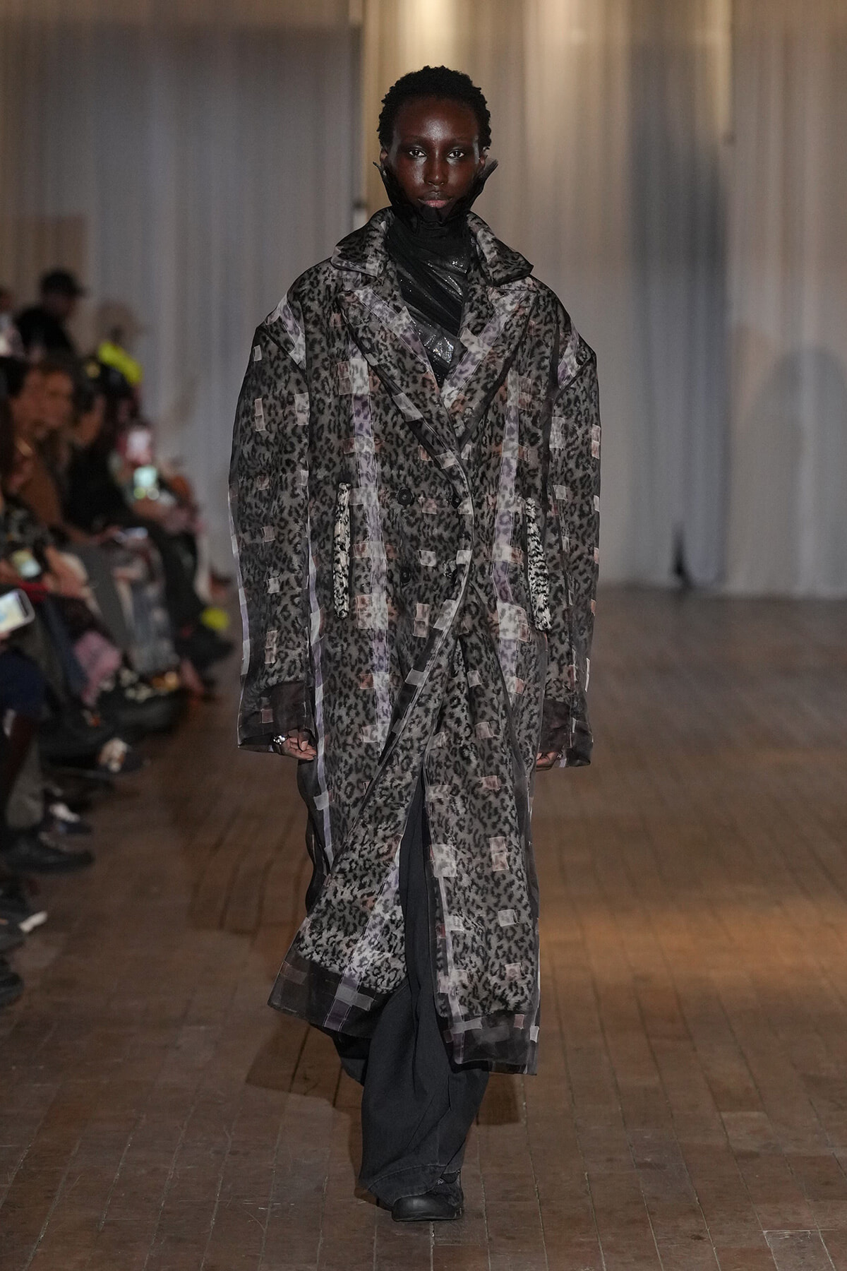 Model walks the runway in a long, oversized leopard-print coat over dark layers, audience in the background.