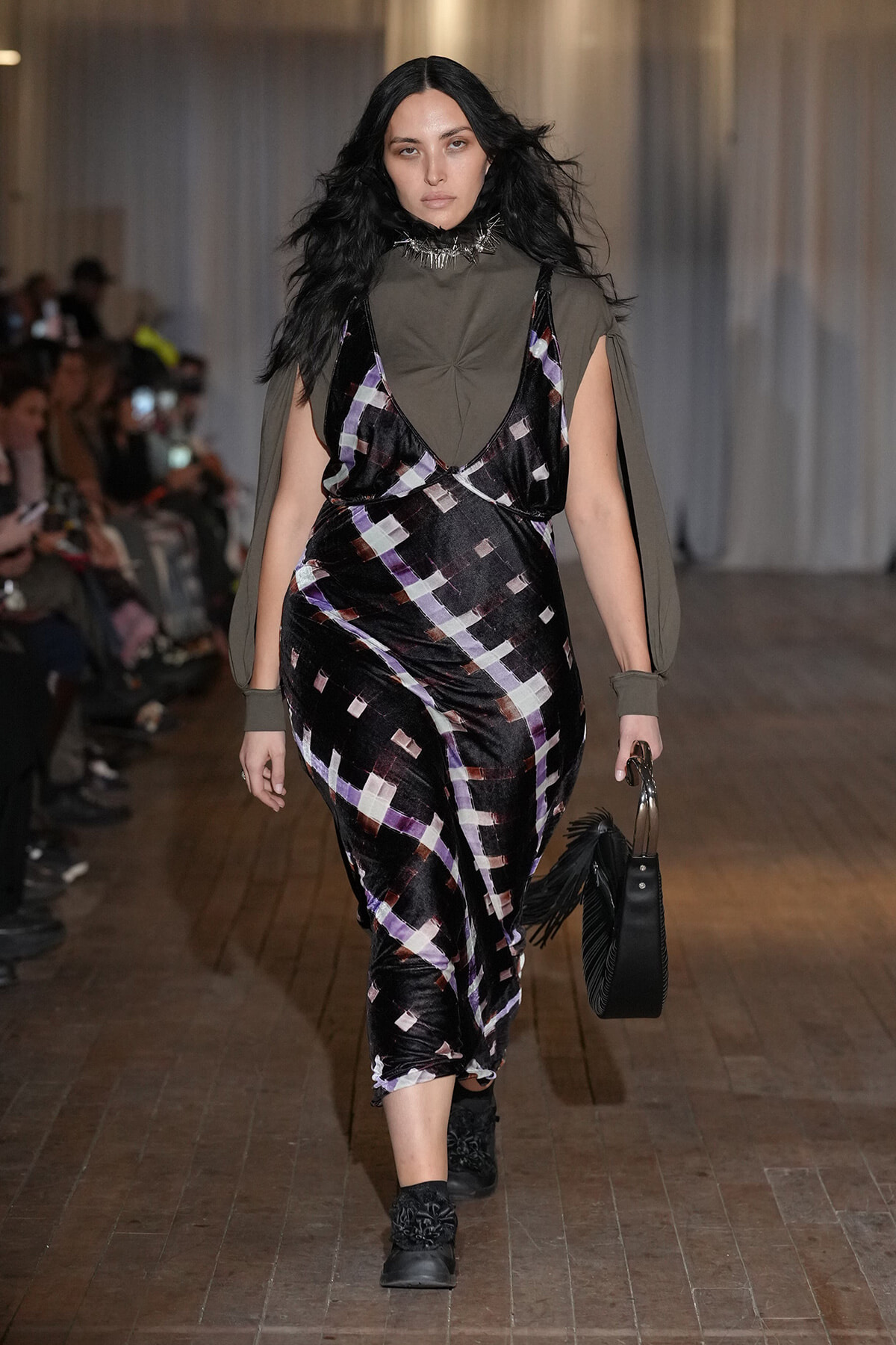 Model walking a fashion show runway in a dark plaid velvet dress layered over an olive-green high-neck top with cape sleeves, carrying a black fringe handbag and wearing black ankle boots.