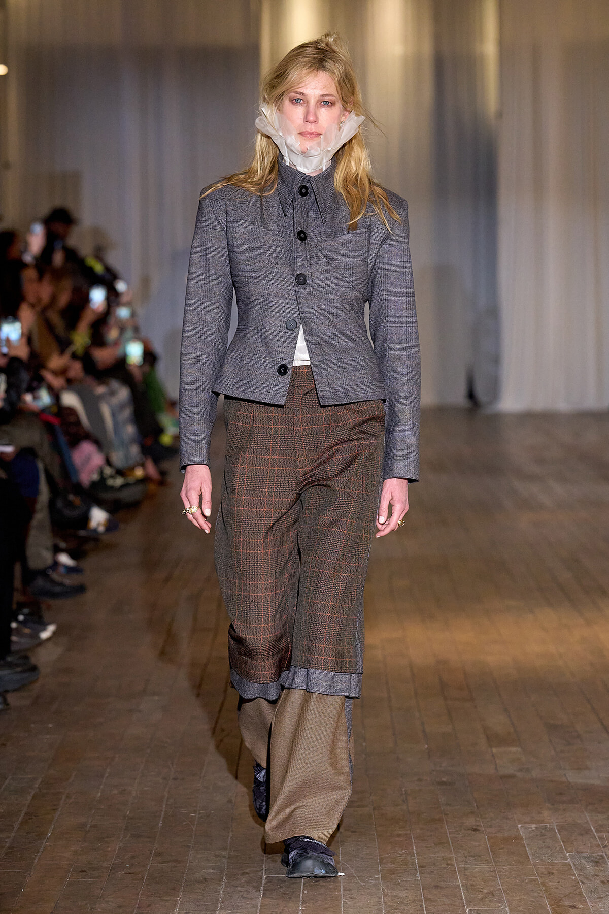 Model on a fashion runway wearing a gray tailored blazer and brown plaid wide-leg pants with cuffed hems, audience in the background