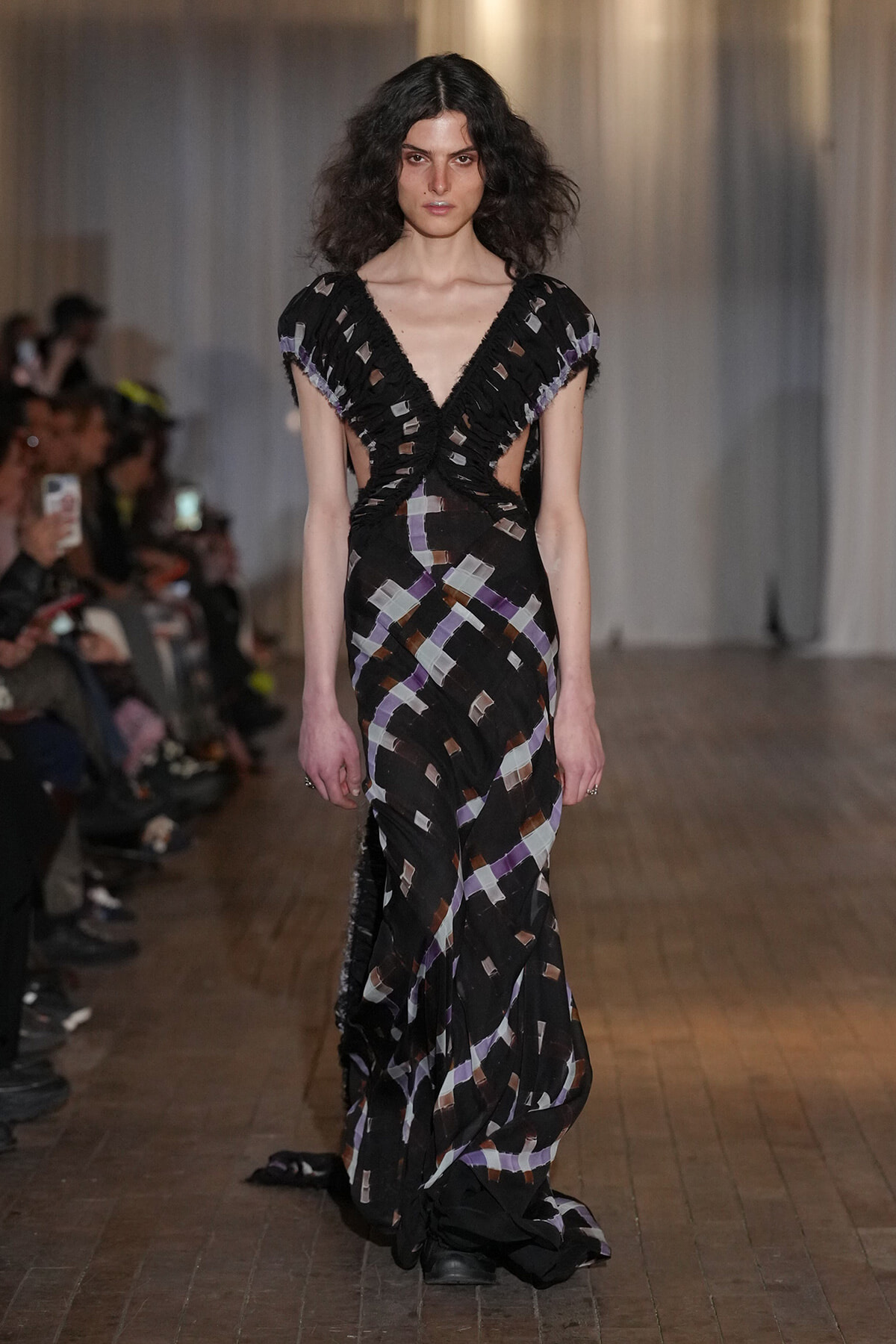 Runway model walking in a black geometric-patterned gown with side cutouts and a plunging V-neck, audience in background