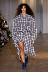 Model on a runway wearing a blue-beige plaid dress with oversized sleeves and cascading ruffles, paired with blue ankle boots.