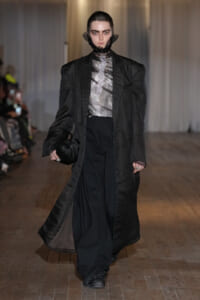 Model walks a runway in a long black coat, sheer patterned top, and wide-leg black pants, carrying a black pleated bag.