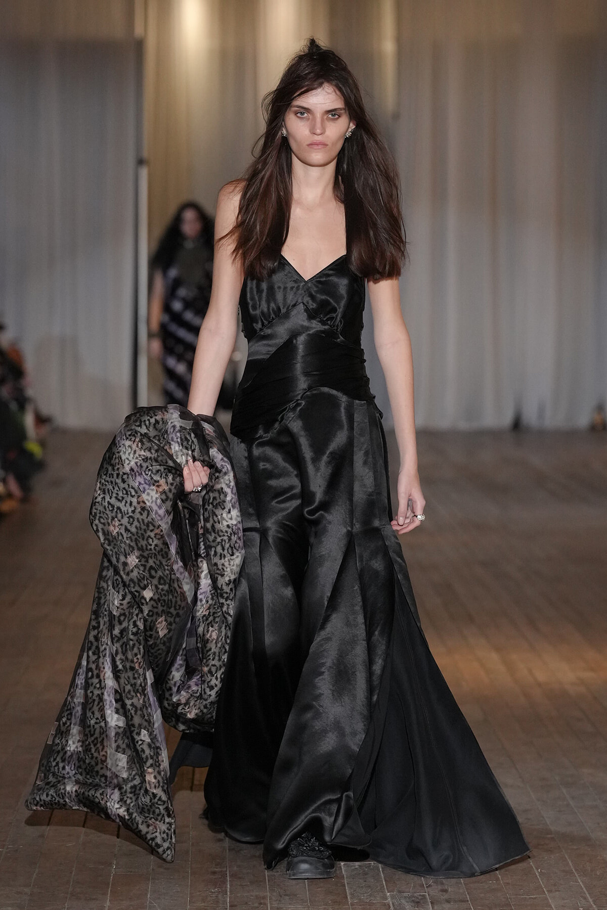 Model walking runway in a black satin gown, holding a leopard-print sheer overlayer in one hand.