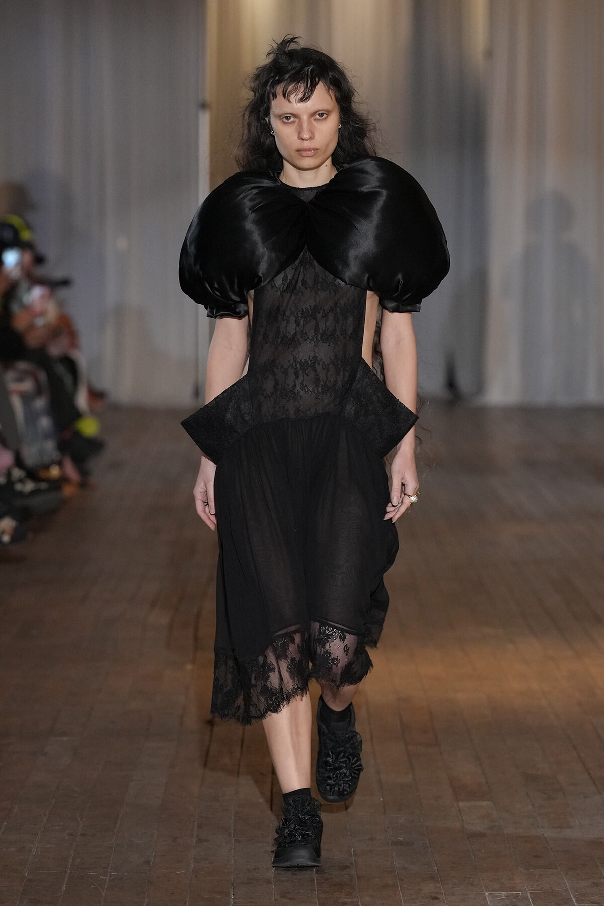 Model walks the runway in a dramatic black ensemble: oversized satin puff shoulders, a lace sheer bodice with a peplum, and a sheer lace-hem skirt paired with black ankle boots.
