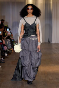 Runway model wearing a layered gray tiered skirt with a black bustier over a beige cape-sleeve top, sunglasses, and a white handbag.