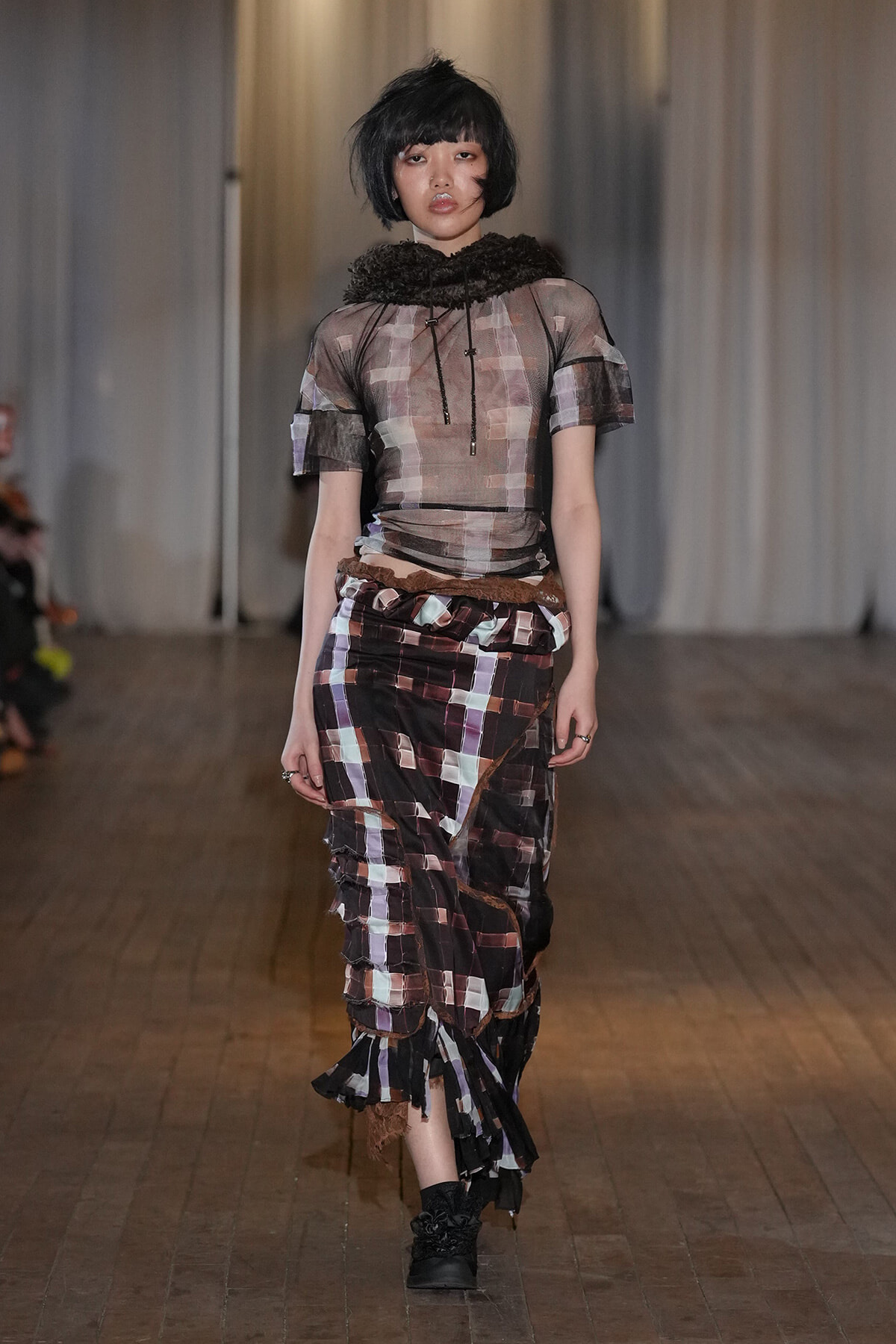 Model walking down a runway in a sheer, patchwork top and multi‑pattern skirt with brown belt and black boots.