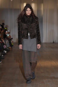 Model walks the runway wearing a black textured blazer with a layered gray plaid trouser ensemble and ankle boots.