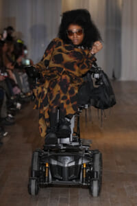 Model in a leopard-print dress on a motorized wheelchair walks a fashion show runway, wearing sunglasses.
