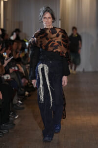 Runway model walking in a leopard-print wrap over a black blouse with navy pinstripe pants and long silver fringe detail at the hip.