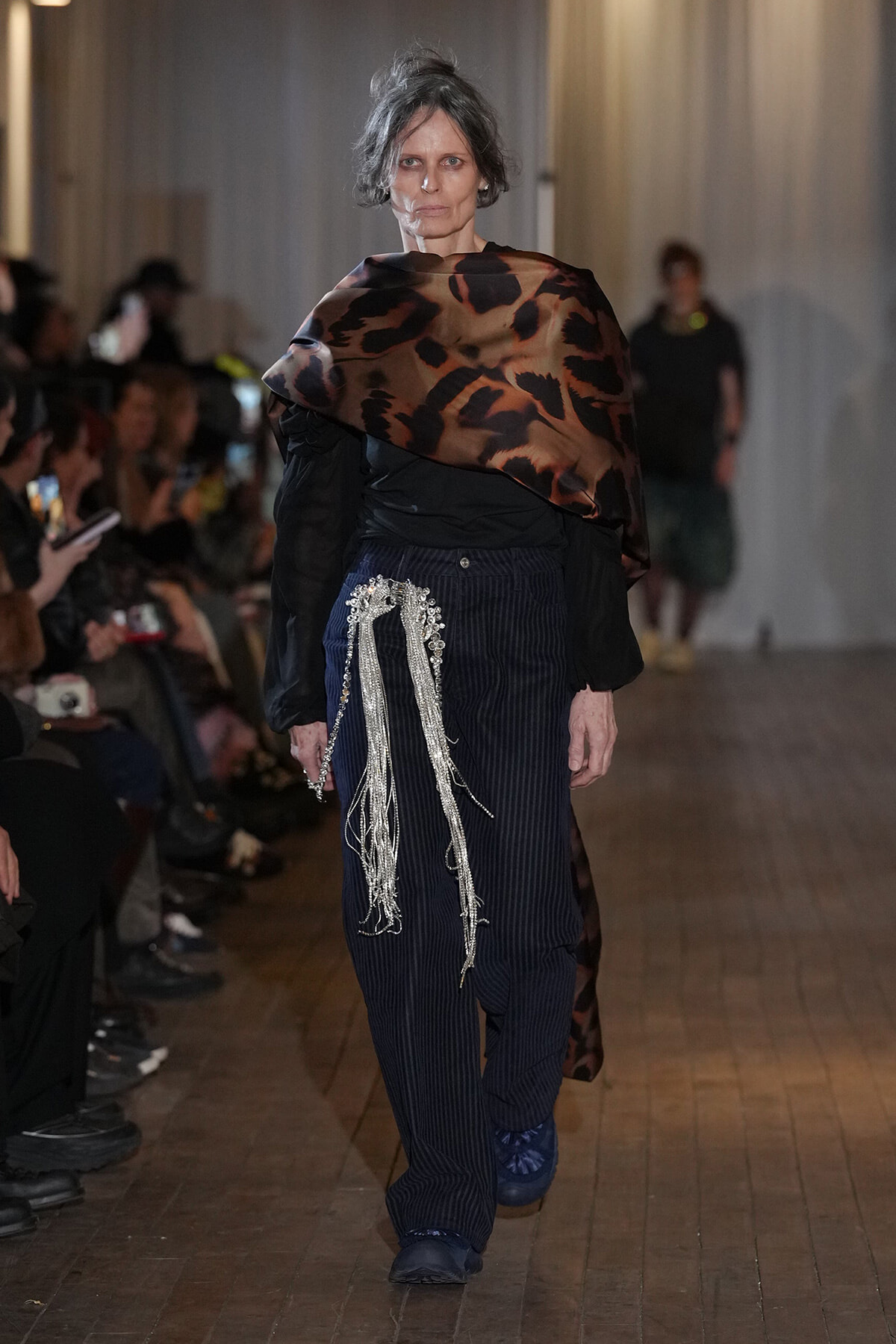 Runway model walking in a leopard-print wrap over a black blouse with navy pinstripe pants and long silver fringe detail at the hip.