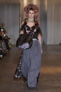 Model walking runway in a high-fashion outfit with oversized gray pants and a black-purple patterned top, carrying a large glossy black bag over her arm.