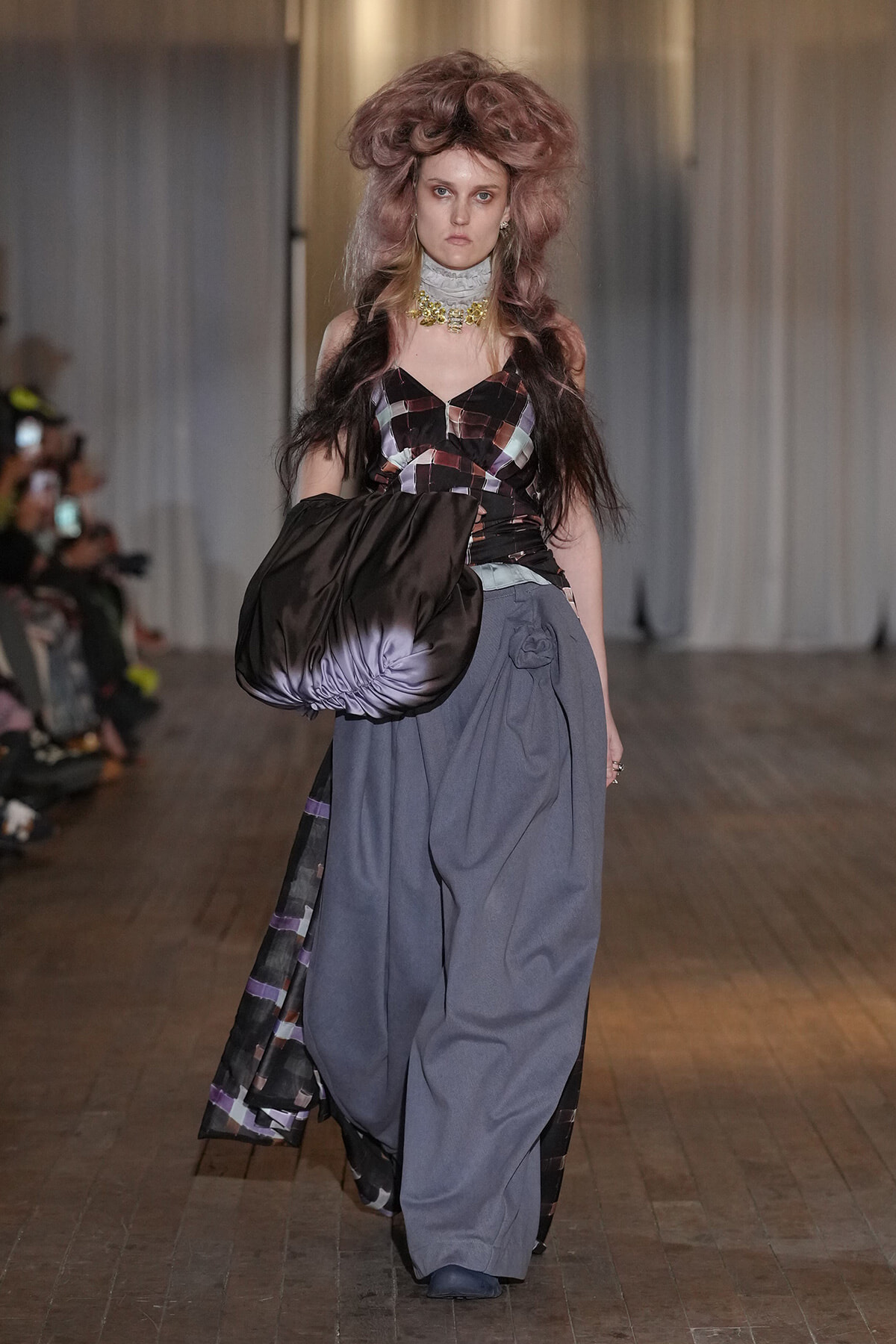Model walking runway in a high-fashion outfit with oversized gray pants and a black-purple patterned top, carrying a large glossy black bag over her arm.