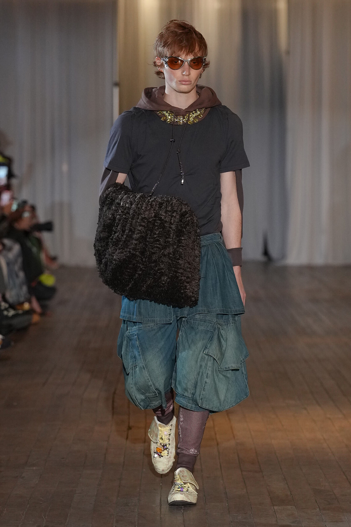 Runway model wearing layered streetwear with oversized teal cargo shorts, dark T‑shirt, brown hoodie, fuzzy black bag, amber sunglasses, and a gold necklace.