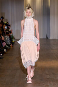 Model walking a runway in a pale pink lace halter top with high ruffle neck and a gathered satin skirt with lace trim at the hem