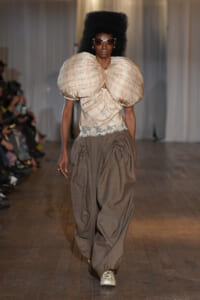 Model walking a runway in an oversized beige top with huge puff sleeves, lace waist, and wide brown draped pants with sneakers, sunglasses, and an afro hairstyle.