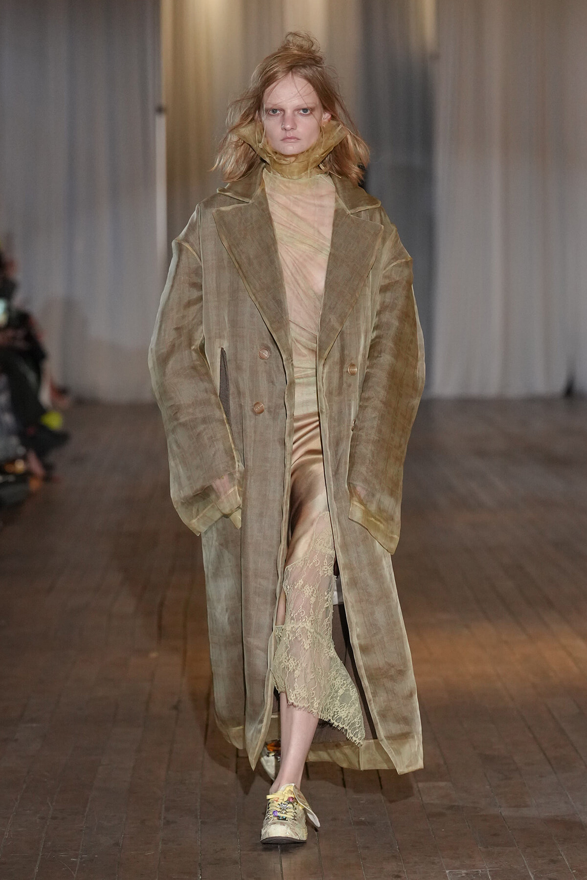 Runway model wearing an oversized beige-brown trench over a sheer champagne dress with lace hem, paired with embroidered sneakers.