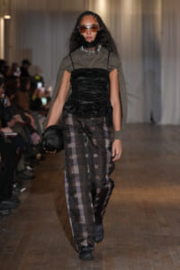 Model walks a fashion runway in a gray cape over a black ruched top and plaid wide-leg pants, wearing orange-tinted sunglasses.