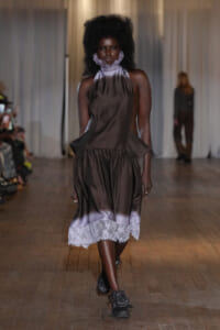 Model walking the runway in a brown satin dress with a white lace hem and a purple high neck scarf, black boots, audience blurred in the background.