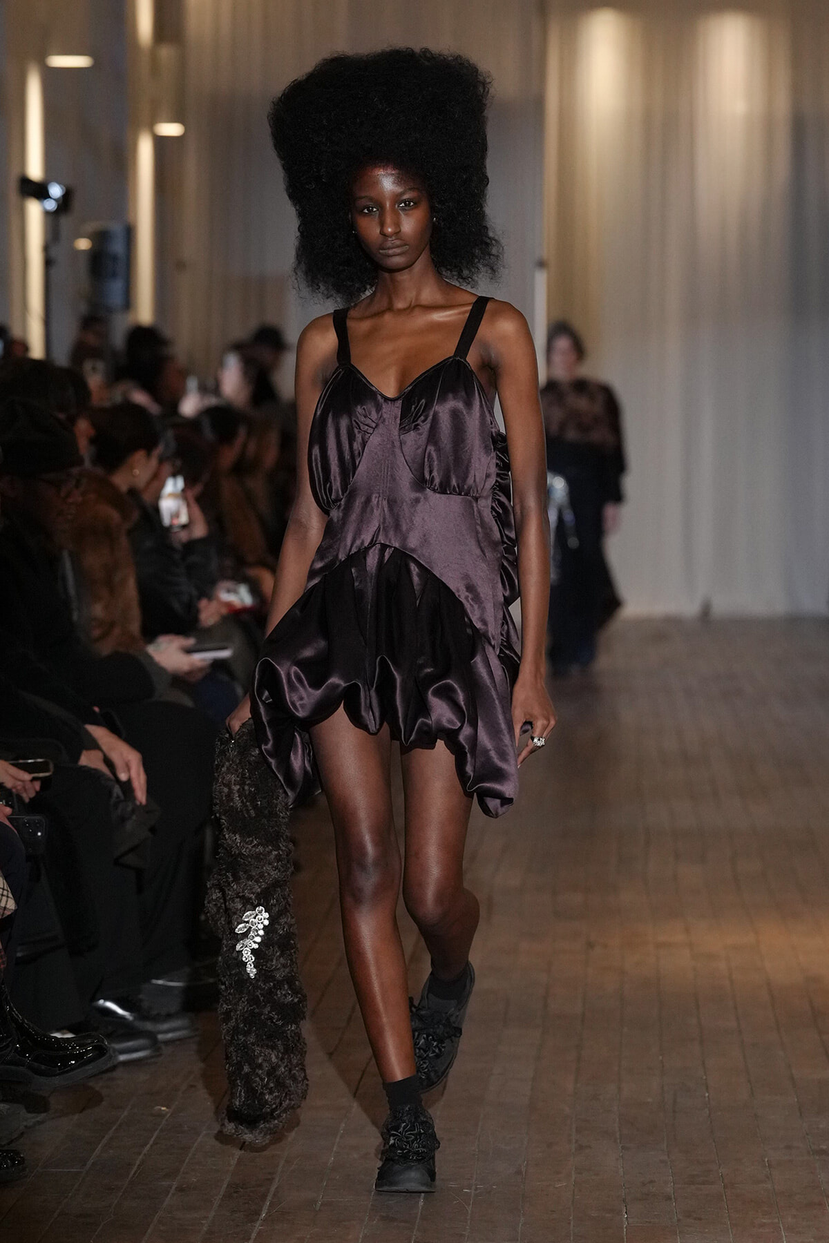 Model walking a runway in a dark purple satin mini dress with a voluminous skirt, holding a textured fur coat in one hand, audience seated along the sides.