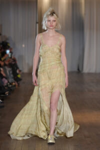 Model walking the runway in a pale yellow, flowing gown with a thigh-high slit and sheer texture.