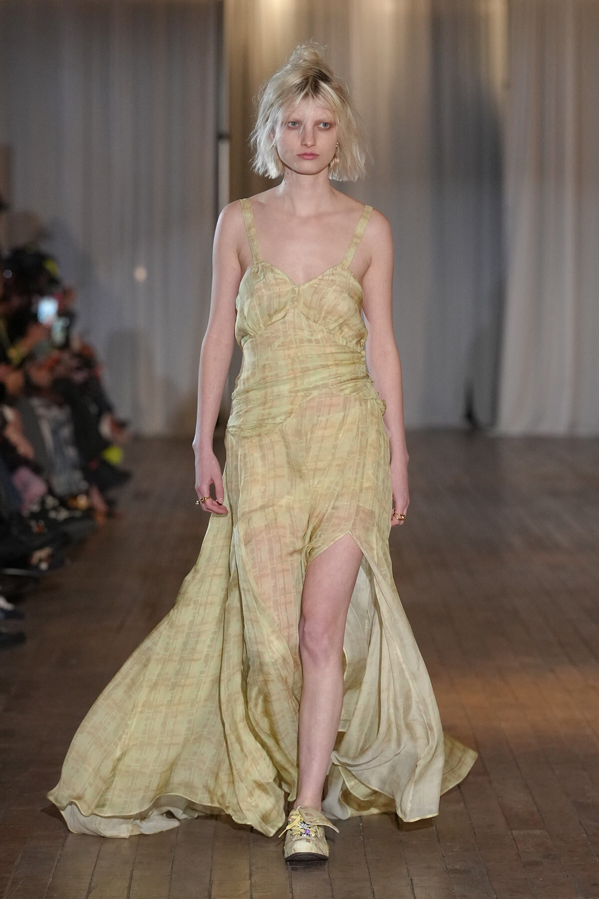 Model walking the runway in a pale yellow, flowing gown with a thigh-high slit and sheer texture.