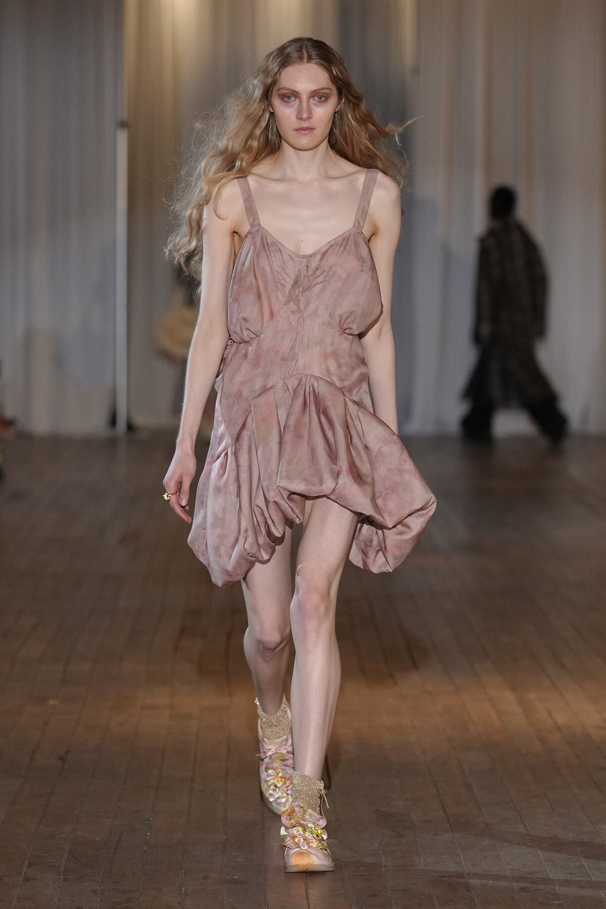 Model walking on runway in a pink, draped sleeveless dress with a voluminous, gathered skirt, neutral makeup and wavy hair.