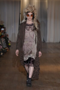 Model walking runway in sheer mauve dress with lace trim, brown blazer, and textured ruffled skirt, orange-tinted sunglasses, voluminous blond hair.