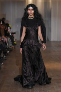 Female model walks a runway in a long black satin gown with layered panels and cape-like sleeves, accessorized with a chunky silver necklace.