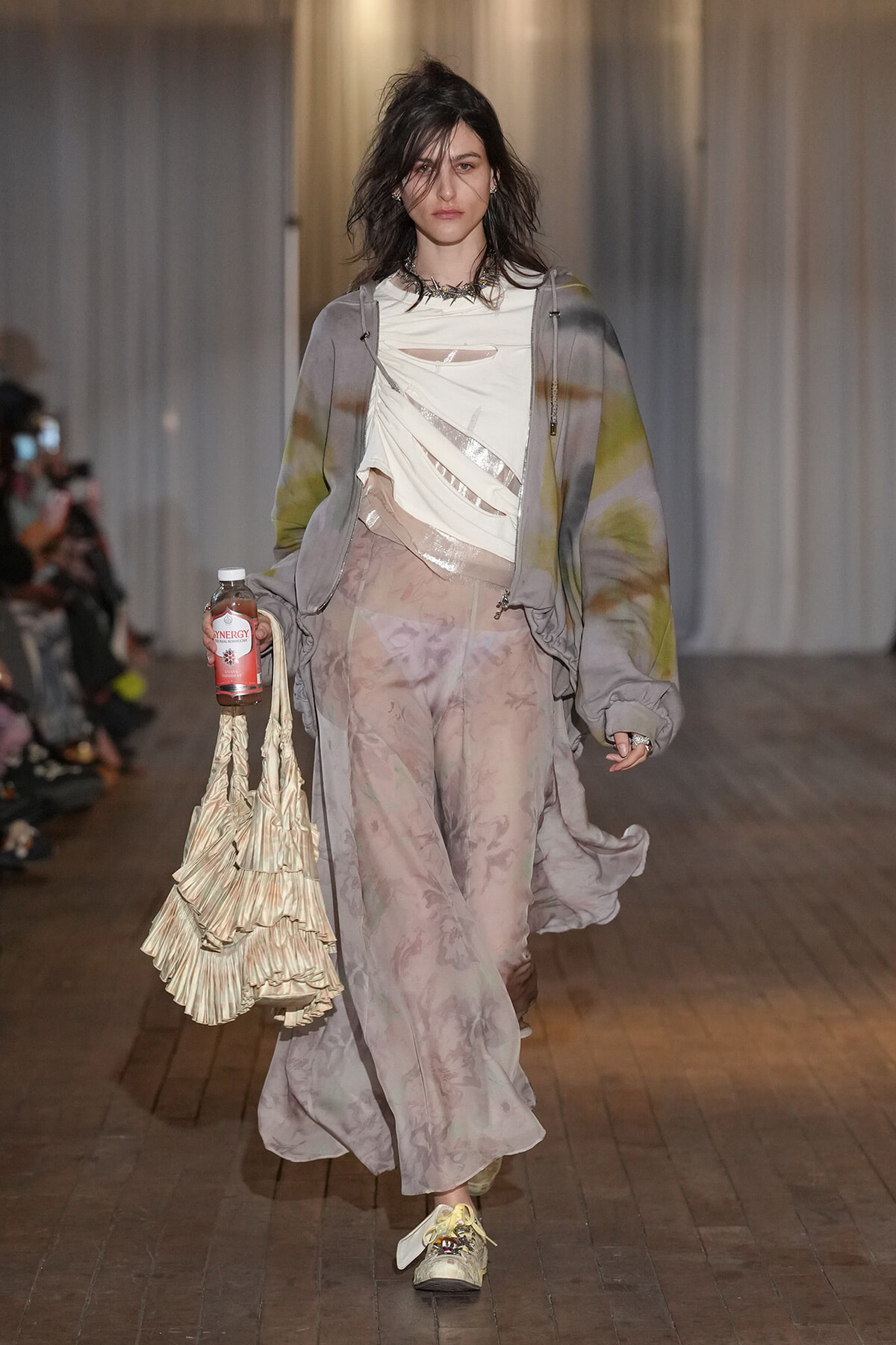 Runway model in a pastel tie-dye oversized hoodie over a twisted cream top and sheer patterned pants, carrying a pleated cream tote and a bottle.