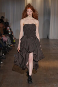 Model walking the runway in a strapless, dark plaid gown with a sculpted bodice and a voluminous, draped skirt.