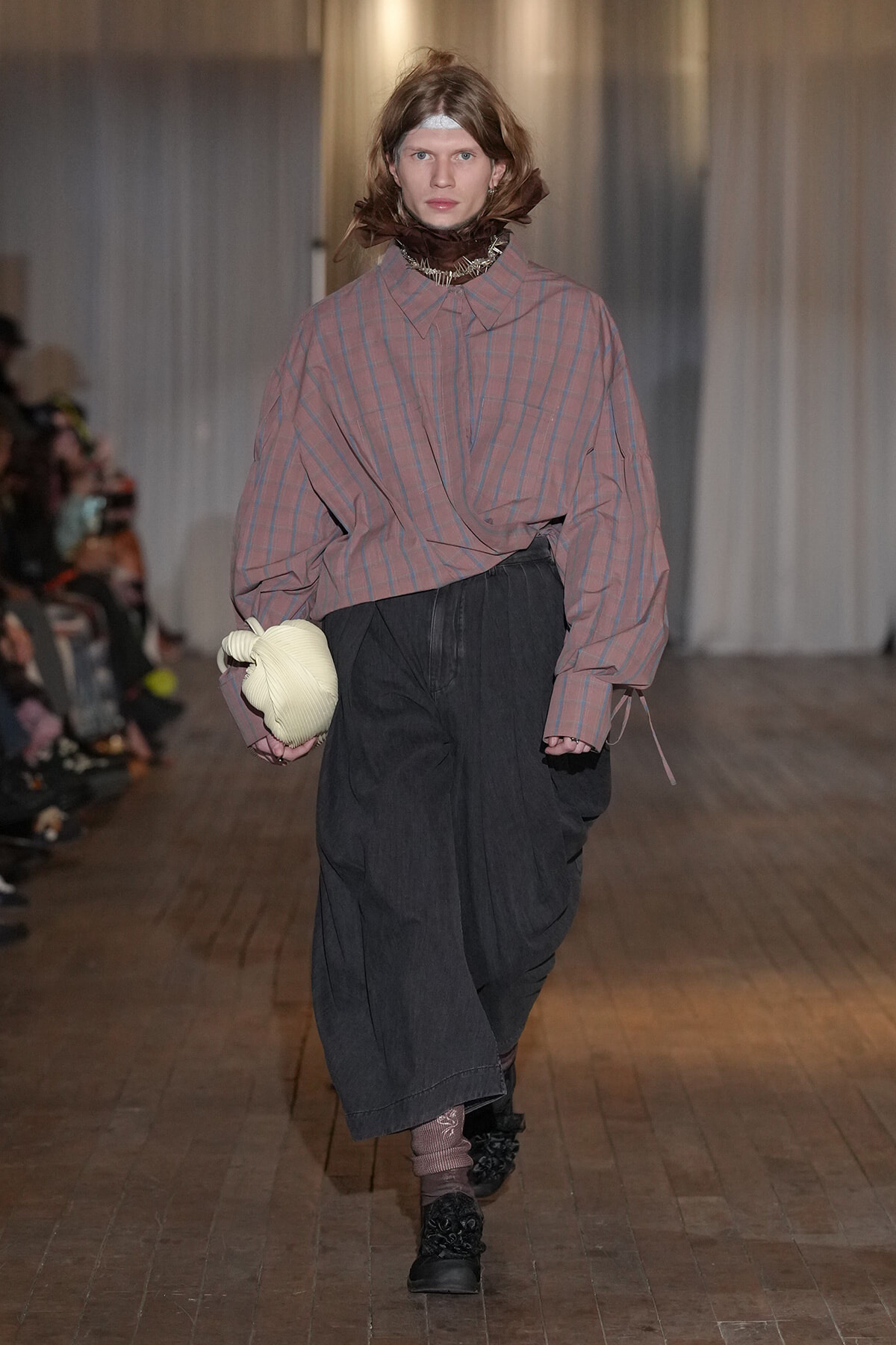 Model on a runway wearing an oversized pink plaid shirt with a high collar, tucked into loose black wide-leg pants; she holds a cream padded clutch.”,