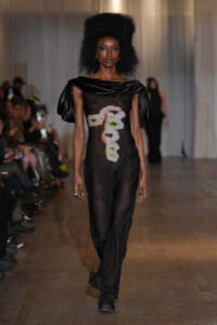 Model walking the runway in a sheer black jumpsuit with oversized off-shoulder sleeves and a vertical floral print down the front, in a dim venue.