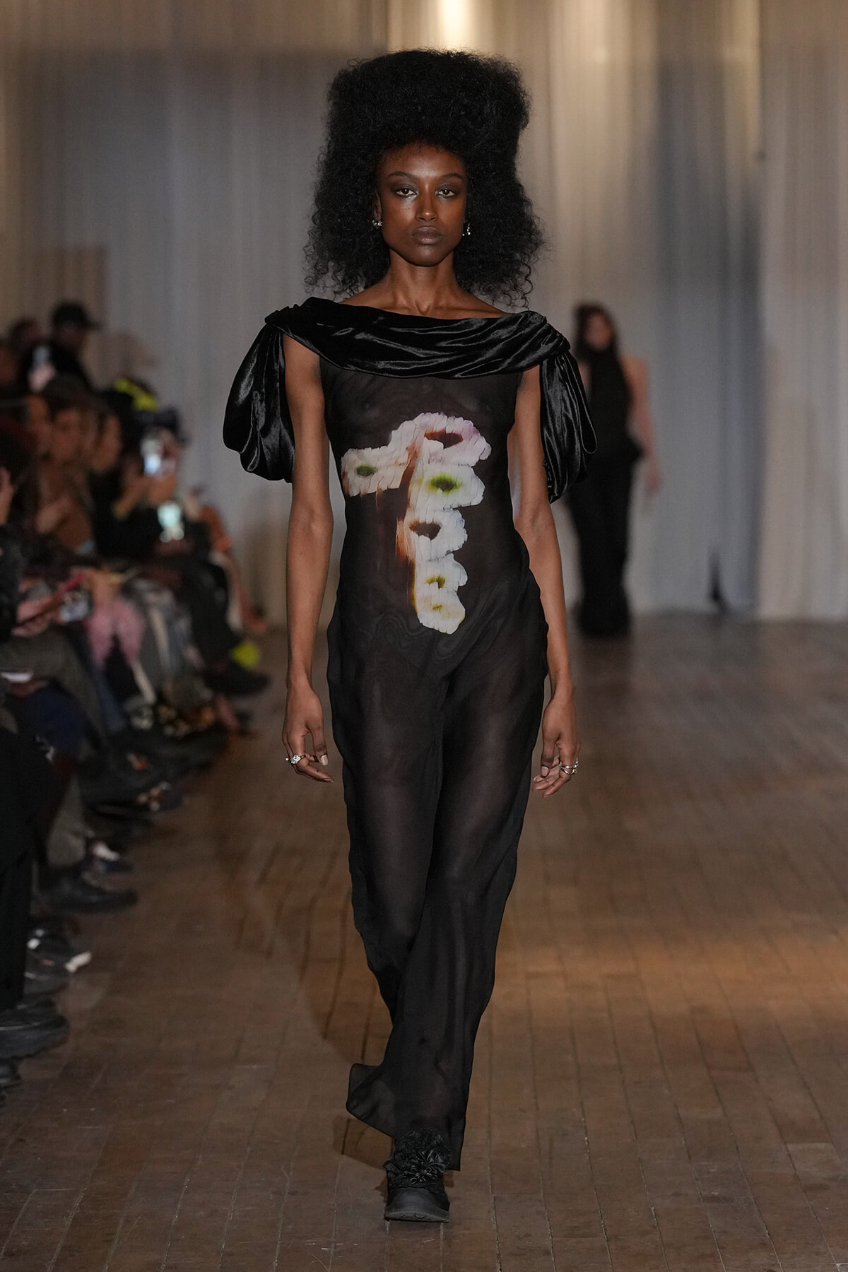 Model walking the runway in a sheer black jumpsuit with oversized off-shoulder sleeves and a vertical floral print down the front, in a dim venue.