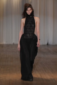 Model walks the runway in a black lace halter top and wide, flowing black pants, on a wooden catwalk.