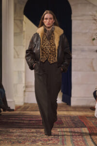 Model on a runway wearing a dark leather jacket with tan shearling, leopard-print top, and wide black trousers