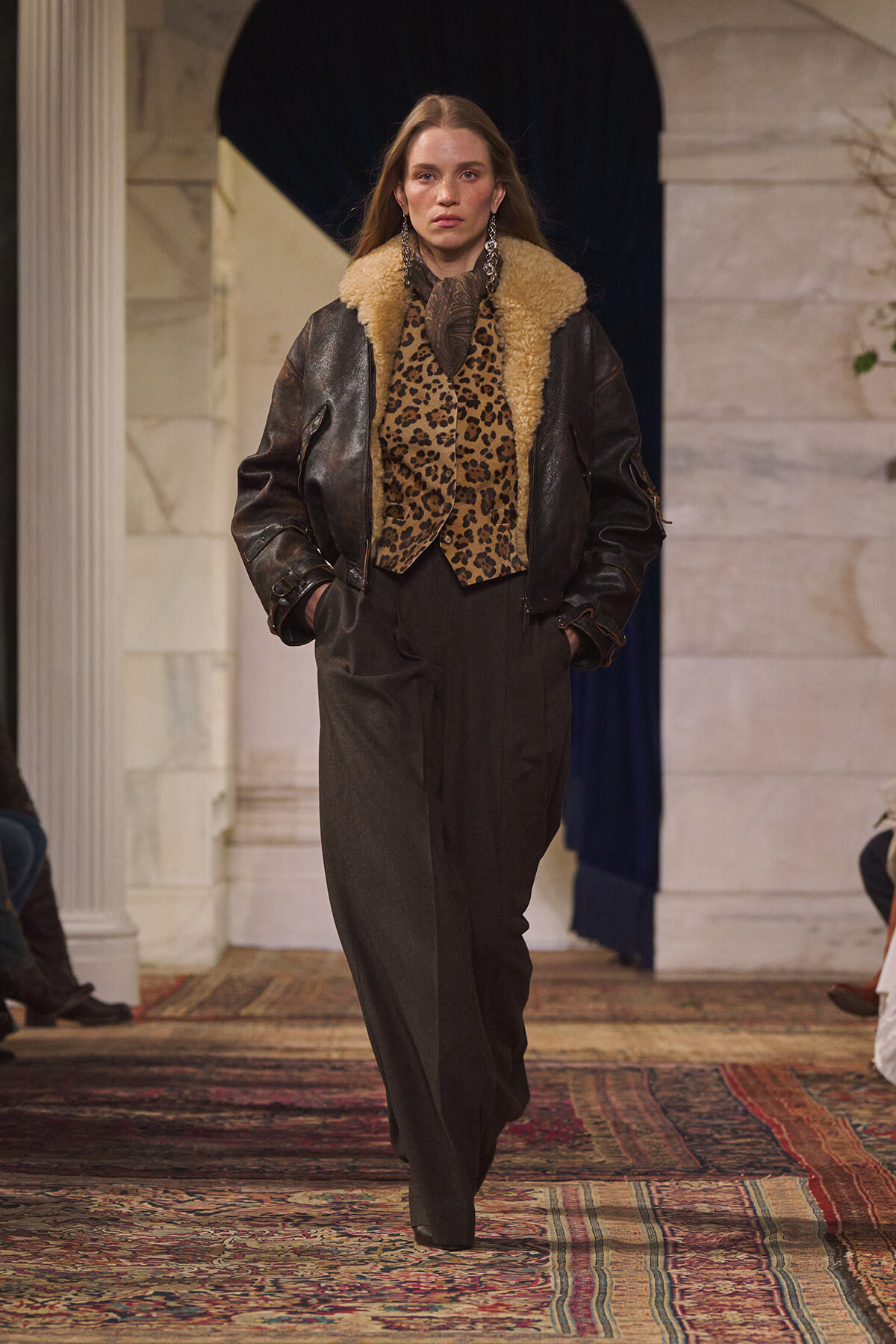 Model on a runway wearing a dark leather jacket with tan shearling, leopard-print top, and wide black trousers
