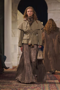 Model on a runway wearing a tan belted trench, metallic pleated skirt, and carrying a beige handbag under arches in a marble setting.