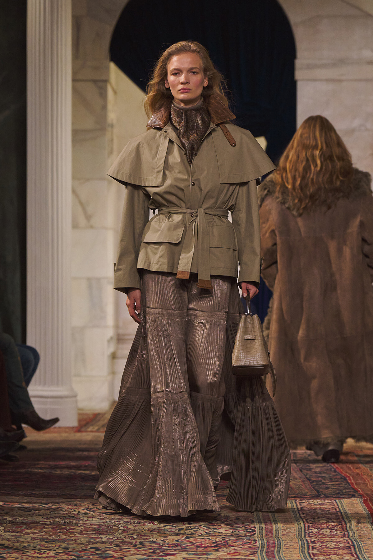 Model on a runway wearing a tan belted trench, metallic pleated skirt, and carrying a beige handbag under arches in a marble setting.