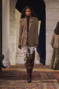 Model walks the runway in a brown tweed blazer over a dark turtleneck, paired with a landscape-print skirt and knee‑high brown boots.