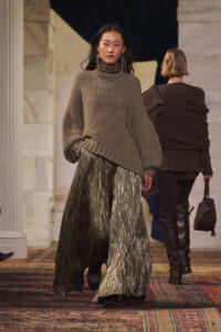 Model walks the runway in an oversized taupe knit sweater paired with a metallic pleated maxi skirt, accessorized with long earrings.