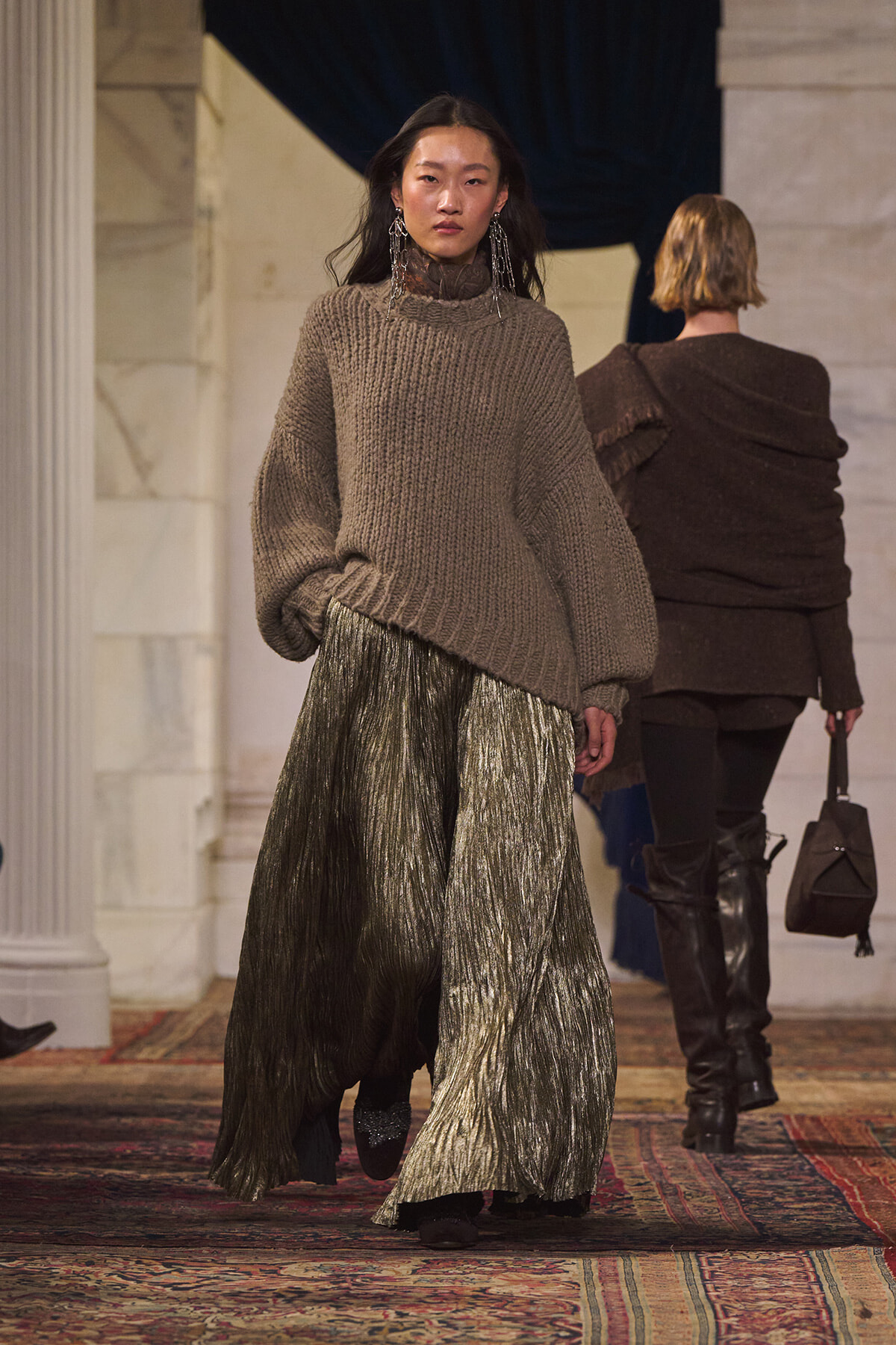 Model walks the runway in an oversized taupe knit sweater paired with a metallic pleated maxi skirt, accessorized with long earrings.