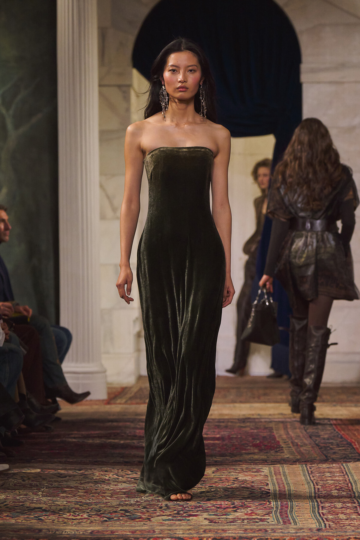 Model walking the runway in a strapless dark green velvet gown with statement earrings.
