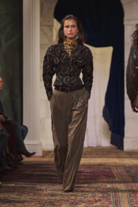 Model on a runway wearing a black lace top, leopard neck scarf, and tan wide-leg pants with a silver chain accessory.