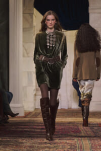 Model walks a fashion runway in a dark green velvet mini dress with a wide brown belt, tall knee-high boots, and bold silver jewelry.