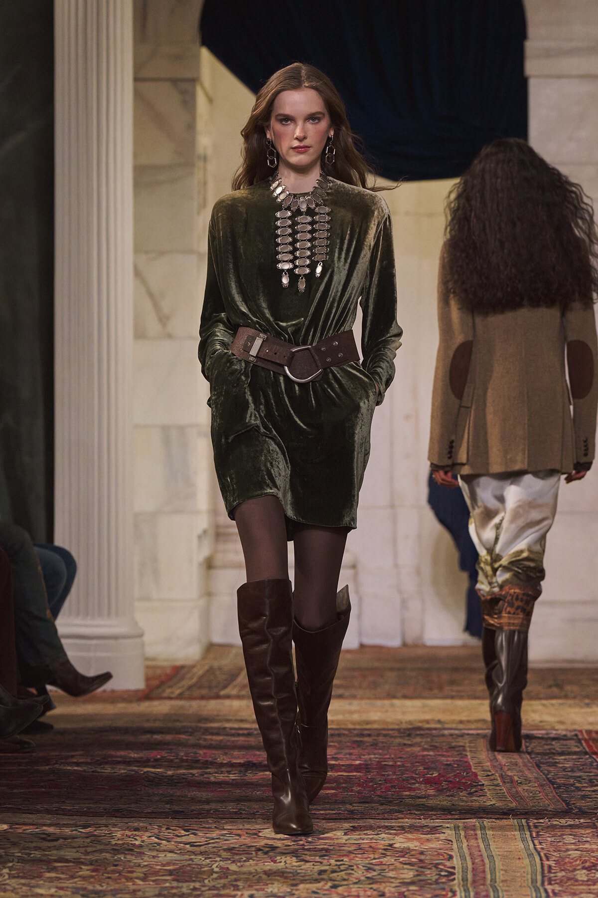 Model walks a fashion runway in a dark green velvet mini dress with a wide brown belt, tall knee-high boots, and bold silver jewelry.